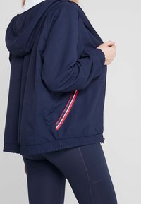 Mörkblå zip-up jacka med huva, som har ett strukturerat tyg, elastiska ärmslut och röda och vita randiga detaljer längs sidorna.