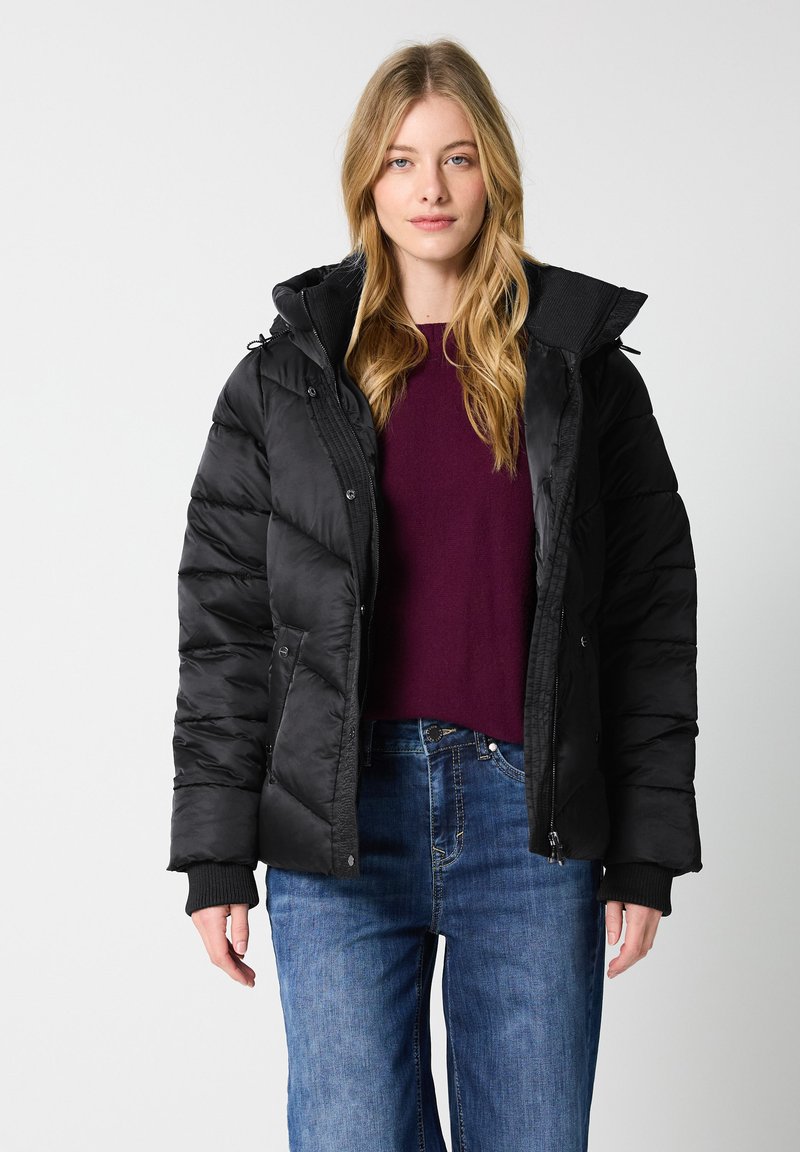 Giacca a vento nera imbottita con zip frontale, cappuccio grande e polsini a coste. Indossata sopra un maglione bordeaux, abbinata a jeans in denim blu.