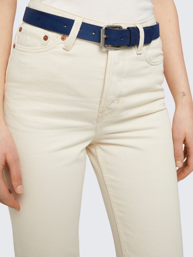 Ceinture en daim bleu marine avec une boucle en métal argenté, portée sur un jean en denim couleur crème avec des rivets en cuivre et une texture subtile.