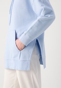 Personne portant un sweat à capuche bleu clair avec des fentes latérales et les mains dans la poche avant, associé à un pantalon côtelé de couleur claire.
