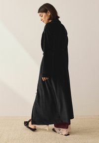 Schwarzer Samt-Langmantel mit Schalkragen, seitlichen Schlitzen und glatter Textur, getragen über einem kastanienbraunen Outfit und mit Spitzenakzenten.