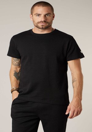 Homme tatoué portant un t-shirt noir uni et un pantalon noir, debout avec une main dans la poche devant un fond neutre.