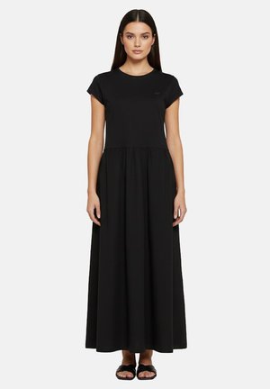 Donna in piedi che indossa un abito maxi nero a maniche corte con vita arricciata e sandali neri open-toe, su uno sfondo bianco uniforme.