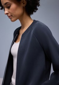 Veste bomber bleu marine foncé avec fermeture éclair à l'avant, col côtelé et texture lisse. Portée sur un débardeur clair, sur un fond neutre.