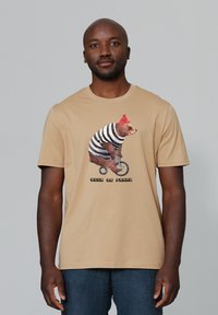 Beige T-Shirt mit einem Bären in einem gestreiften Anzug und rotem Hut, der Fahrrad fährt. Darunter steht "Pour le Franz." Aus Baumwolle, lässige Passform.