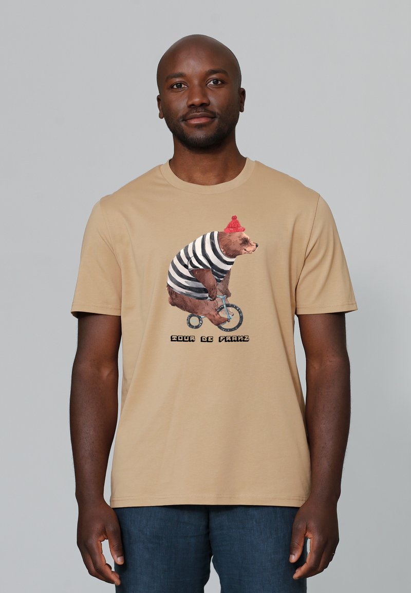 Beige T-Shirt mit einem Bären in einem gestreiften Anzug und rotem Hut, der Fahrrad fährt. Darunter steht "Pour le Franz." Aus Baumwolle, lässige Passform.