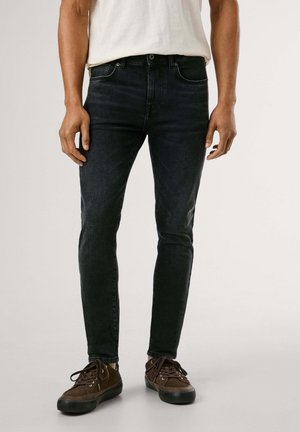 Homme portant un jean noir slim, un t-shirt blanc et des baskets marron à lacets, debout devant un fond clair uni.