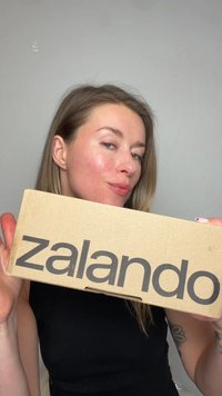 Pitkähiuksinen nainen pitää kasvojaan lähellä harmaata taustaa vasten olevaa „Zalando”-tekstillä varustettua pahvilaatikkoa.