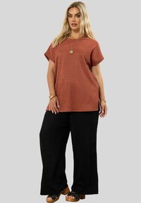 Yläosa ruosteenvärisiä tekstuureja, lyhythihaisena, yhdistetty mustiin leveisiin housuihin. Huomattavat sivuaukot yläosassa ja avonaiset kengät puupohjalla.