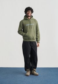 Olivgrüner Kapuzenpullover mit Fronttasche und blauem Logo, kombiniert mit schwarzen Cargo-Hosen und beigen Stiefeln auf einem blauen Teppich.