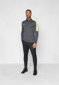 Grijze langemouwen sporttop met neon groene schouderpanelen, gecombineerd met zwarte strakke broek en zwarte sneakers. Nike-logo's op de top en broek.