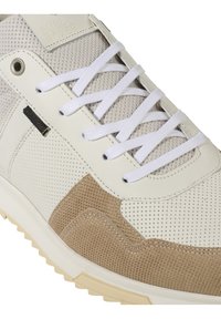 Zapatilla blanca y beige que combina materiales de malla y gamuza, textura perforada, cordones blancos y una suela de goma. Logo de la marca cerca de la parte superior.