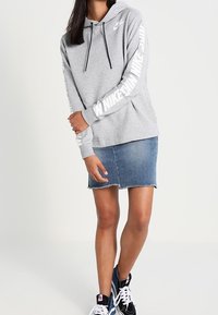 Šedá mikina s kapucí a šňůrkami, s bílým textem "Nike" podél rukávů. Nositá přes džínovou sukni a doplněná černými teniskami.