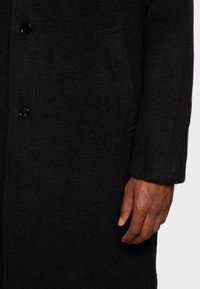 Schwarzer Wollmantel mit glatter Textur, zwei vorderen Knöpfen und seitlicher Tasche, in leicht übergroßer Passform und mit umgeschlagenen Ärmeln.