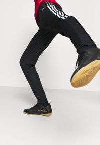 Black athletic pants with white side stripes, featuring a slim fit. Worn with black shoes having yellow accents, highlighted by the word "NEMESIS."