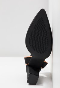 Chaussure noire à talons hauts avec un bout pointu, semelle en caoutchouc texturée, et un accent orange sur la sangle de la cheville. Présente un design de talon effilé.