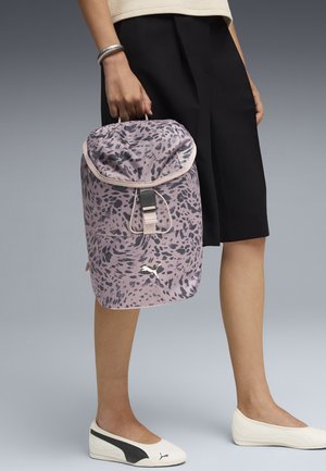 Personne tenant un sac à dos Puma violet à pois noirs par la poignée supérieure, portant une jupe noire et des chaussures Puma blanches sans lacets.