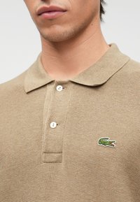 Μπεζ πόλο μπλουζάκι σε υφασμάτινη πλέξη με ριγωτό γιακά, διαθέτει δύο λευκά κουμπιά και ένα μικρό πράσινο λογότυπο κροκοδείλου στο στήθος.