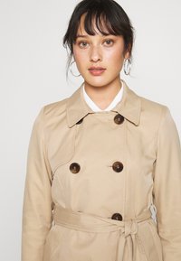 Bežový trench coat s dvouvrstvým designem, velkými tmavými knoflíky a zav�ázaným páskem v pase. Hladká textura, klasický límec.