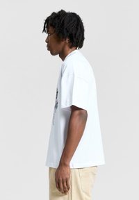 Valkoinen puuvillapaita lyhyillä hihoilla, jossa on värikäs graafinen printti etupuolella. Suunnittelussa on rento ja ylisuuri muoto.