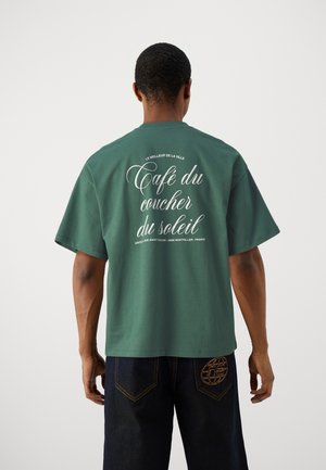 Grünes Baumwoll-T-Shirt mit kurzen Ärmeln und Rundhalsausschnitt. Auf der Rückseite befindet sich weißer Text: "Café du coucher du soleil" sowie ein Logo an der Seite.