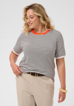 Frau mit blondem lockigem Haar, die ein schwarz-weiß gestreiftes Shirt mit einem orangefarbenen Kragen, beige Hose, einen braunen Gürtel und ein beiges Armband trägt.