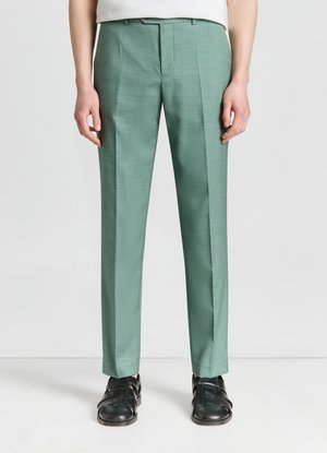 Uomo che indossa pantaloni su misura verde chiaro slim fit e sandali in pelle neri, in piedi davanti a uno sfondo bianco semplice.