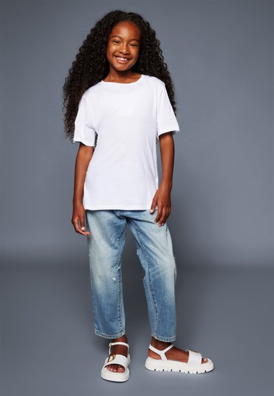 T-shirt bianca a maniche corte, jeans chiari strappati, sandali bianchi con accento dorato; capelli ricci, sfondo neutro.