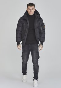 Schwarze Pufferjacke mit Kapuze, ausgestattet mit gerippten Bündchen und einer Seitentasche. Trägt ein schwarzes Rollkragenoberteil und kombiniert mit dunklen Cargo-Jeans und weißen Sneakers.