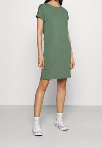 Zielona bawełniana sukienka w kształcie shift z krótkimi rolowanymi rękawami, bocznymi rozcięciami oraz prostym okrągłym dekoltem. W połączeniu z białymi sneakersami typu high-top.