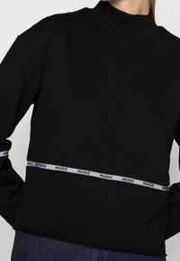 Osoba oblečená v černém tričku s dlouhými rukávy, které má bílý horizontální pruh se opakujícím se textem "HUGO" na střední části a rukávech.