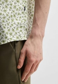 Camicia a maniche corte in tessuto bianco con motivo di fogliame verde; etichetta del logo visibile. Abbinata a pantaloni verde oliva. Mano appoggiata di lato.