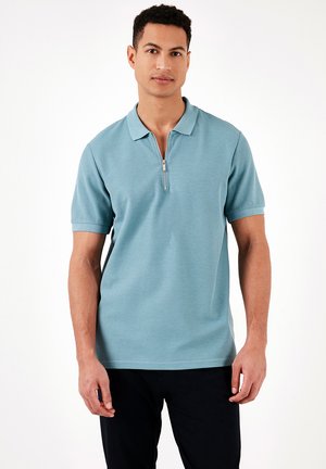 Polo di colore azzurro chiaro con tessuto a trama, maniche corte e colletto con mezza zip. Indossato con pantaloni scuri; nessun motivo o accento visibile.