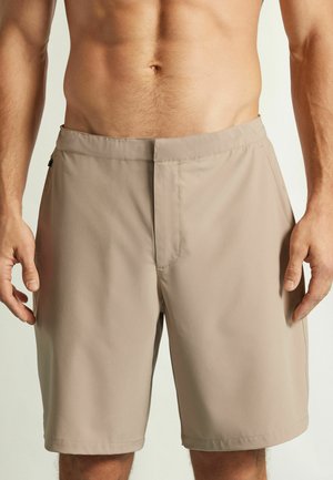 Beige Herren-Knielange Shorts mit flacher Vorderseite und Seitentaschen, getragen von einer oberkörperfreien Person vor einfarbigem Hintergrund.