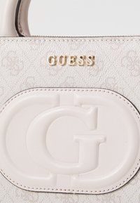 Gjett håndveske i lys beige teksturert stoff med preget logo. Har detaljer i gullfarget metall og et glatt læraccent. Glidelås-lukking.