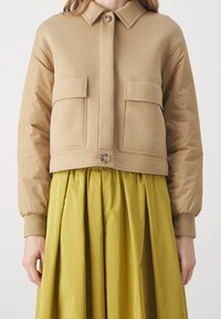 Beżowa krótka kurtka z kołnierzykiem zapinanym na guziki, z dwoma przednimi kieszeniami i ściągaczami przy mankietach, nałożona na żółtą spódnicę z zakładkami.