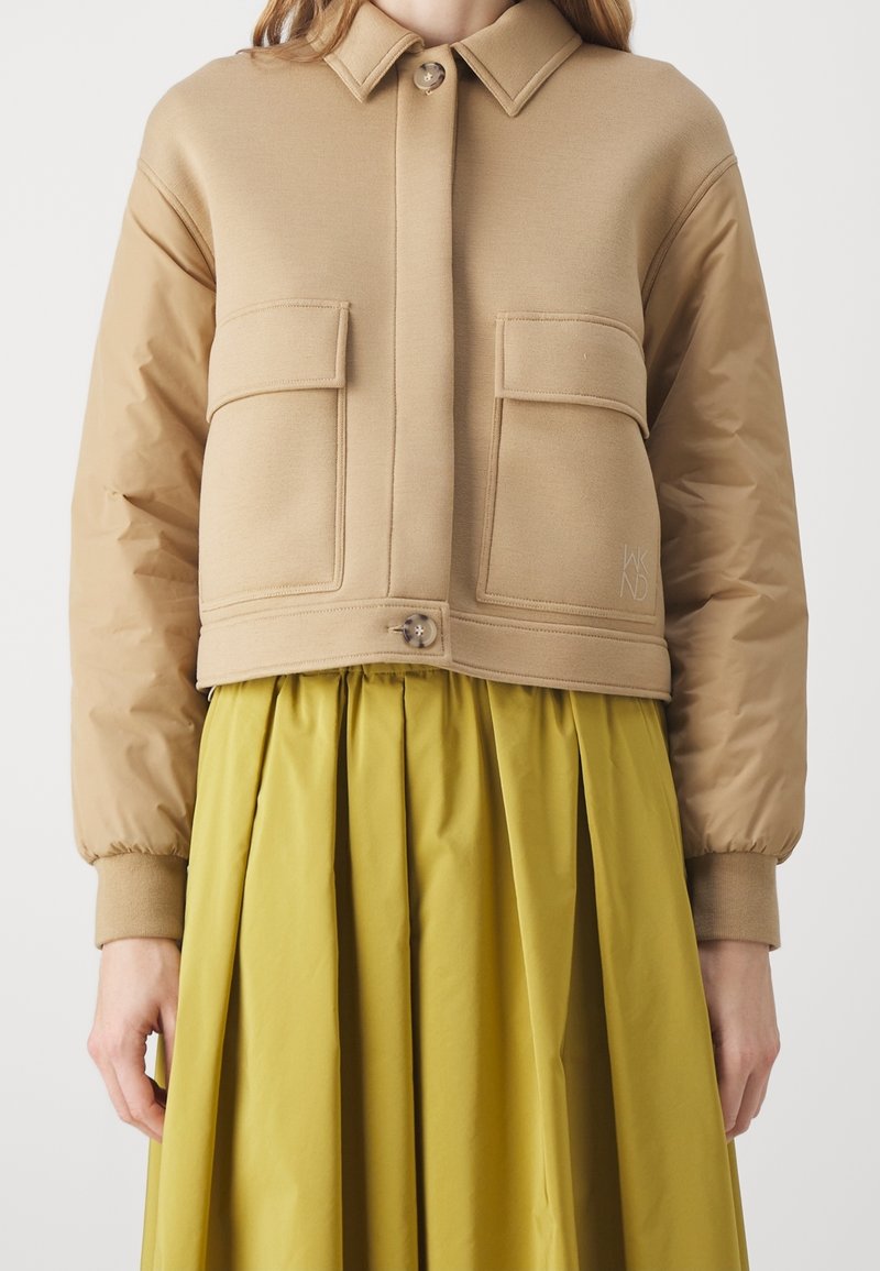 Beżowa krótka kurtka z kołnierzykiem zapinanym na guziki, z dwoma przednimi kieszeniami i ściągaczami przy mankietach, nałożona na żółtą spódnicę z zakładkami.