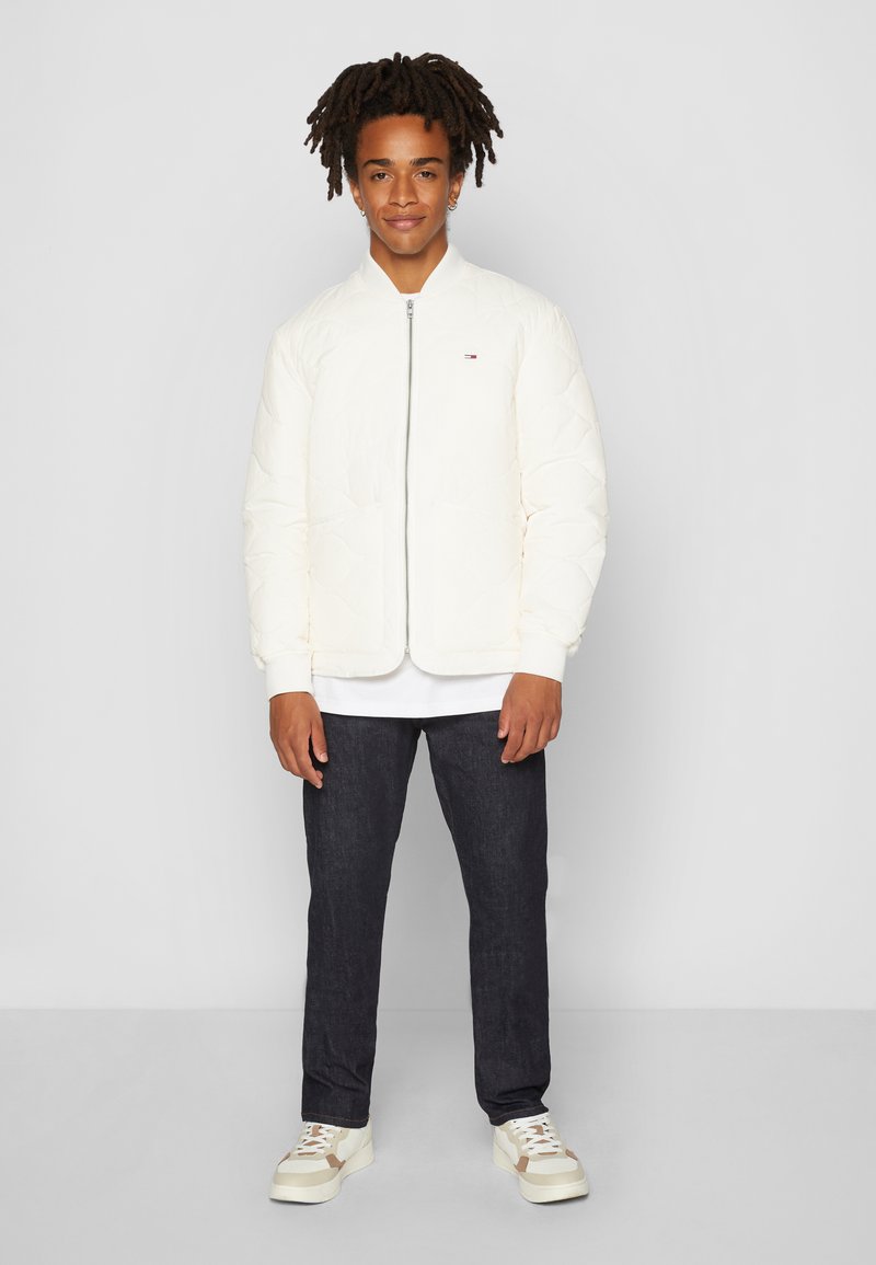 Witte gewatteerde bomberjack met een voorkantoor, ribgebreide mouwen en een klein logo. Draagt over een wit shirt, gecombineerd met donkere jeans en witte sneakers.