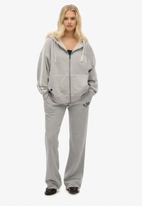 Mujer rubia de pie con las manos en los bolsillos, vistiendo una sudadera gris con cremallera y pantalones de chándal a juego, con zapatos oscuros, sobre un fondo blanco.