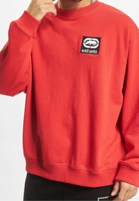 Rode crewneck sweatshirt van katoen, met een zwart rechthoekig logopatch met witte tekst en ontwerp op de linkerkant van de borst.