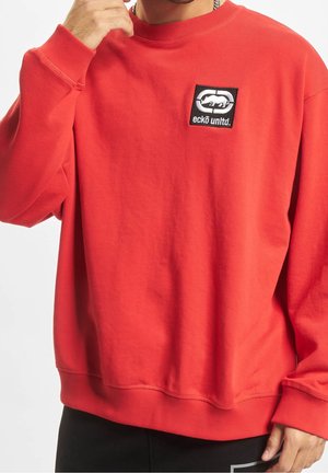Sudadera roja de cuello redondo hecha de algodón, que presenta un parche de logo rectangular negro con texto y diseño en blanco en el lado izquierdo del pecho.