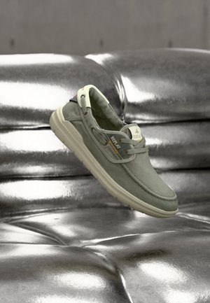 Καθημερινό γκρι υφασμάτινο παπούτσι με λευκή σόλα και την επωνυμία "Replay", αιωρούμενο πάνω από μια λαμπερή ασημί δερμάτινη καναπέ με καπιτονέ.