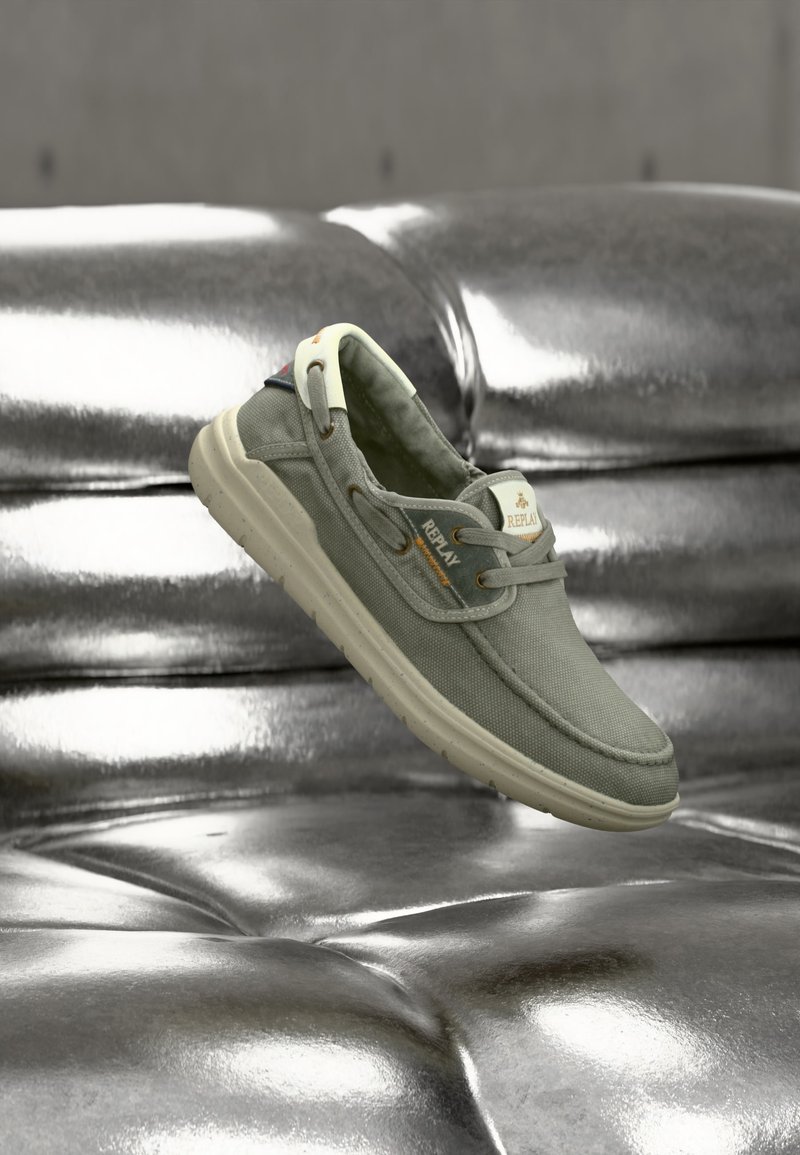 Καθημερινό γκρι υφασμάτινο παπούτσι με λευκή σόλα και την επωνυμία "Replay", αιωρούμενο πάνω από μια λαμπερή ασημί δερμάτινη καναπέ με καπιτονέ.