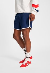 Marinblå sportshorts med vita kantband, kombinerade med vita strumpor med svarta chevronmönster, och röda sneakers med vita detaljer.