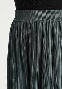 Falda plisada de color verde oscuro con una ancha cintura elástica. La tela tiene un suave brillo y rayas verticales que recorren toda la textura.