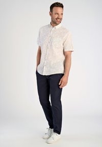 Camicia a maniche corte con bottoni di colore crema con un leggero motivo, abbinata a pantaloni blu navy e sneakers bianche. Presenta una tasca sul petto e una texture liscia.