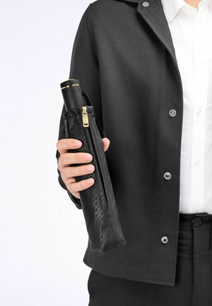 Ombrello nero in un rivestimento nero testurizzato con zip. Il manico è accentuato in oro e l'outfit è scuro e aerodinamico.