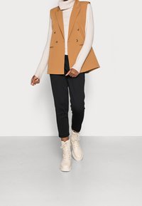 Velbloudí dvouřadý svetr s rozšířeným designem, spárovaný s krémovým rolákem a černými zúženými kalhotami. Krémové kotníkové boty na šněrování.