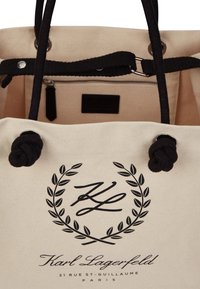 Beige canvas tote bag met zwarte touwhandvatten, met een logodesign en een ritsvak aan de binnenkant. Metalen accenten aan de bovenrand.