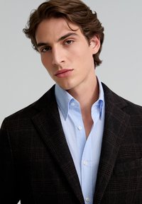 Blazer marrón a cuadros sobre una camisa de dress de rayas azul claro. El blazer presenta solapas de muesca y una tela texturizada. Se ven botones en la camisa.